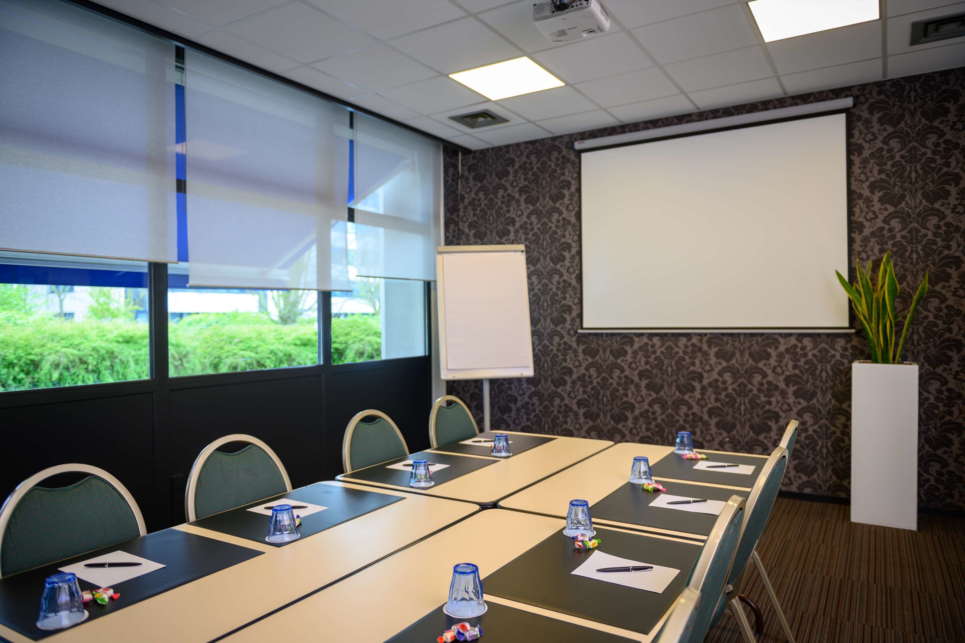 Un salón de conferencias con una larga mesa, sillas, una pantalla de proyector y una planta en maceta.