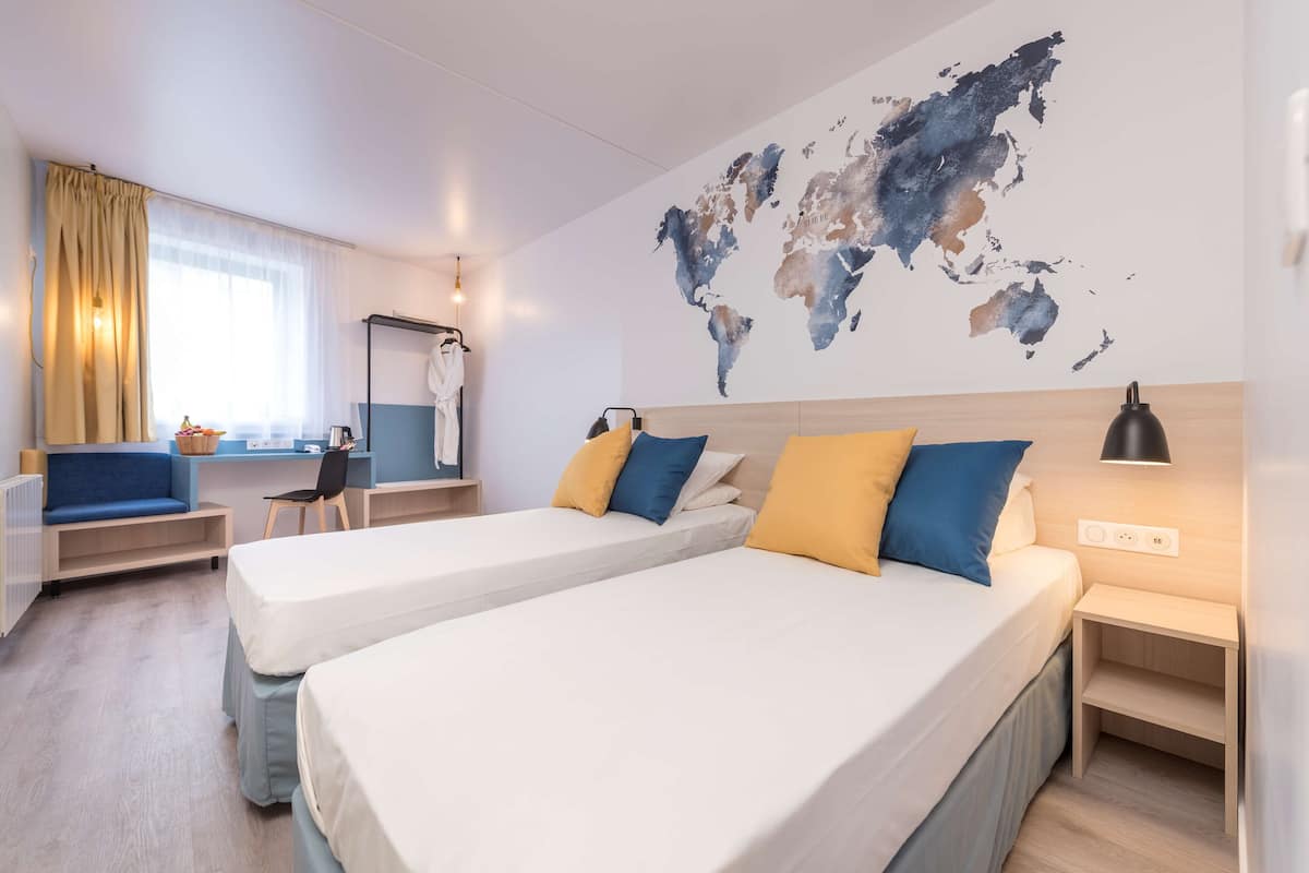 Une chambre d’hôtel moderne avec deux lits, une carte du monde accrochée au mur, un bureau avec une chaise et une fenêtre avec des rideaux.