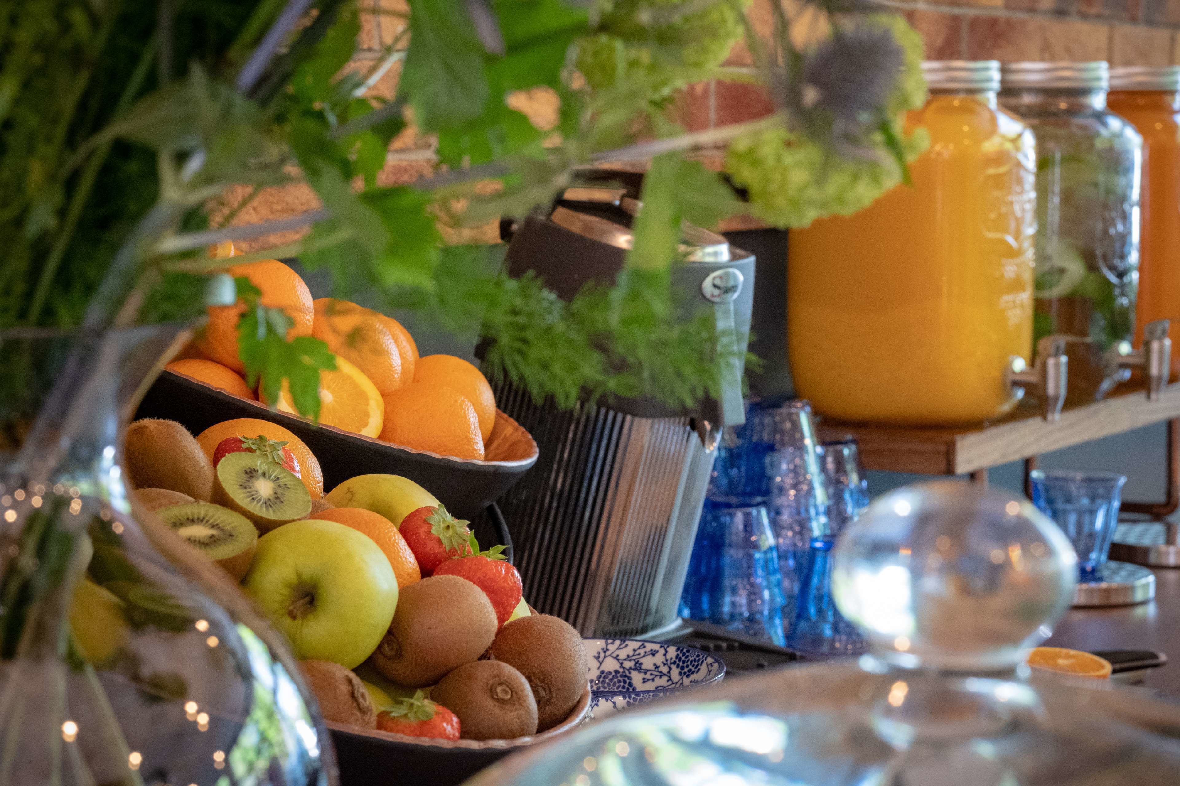 A table with a fruit bowl containing kiwis, apples, oranges, and strawberries, a large orange jug, and a metal container.