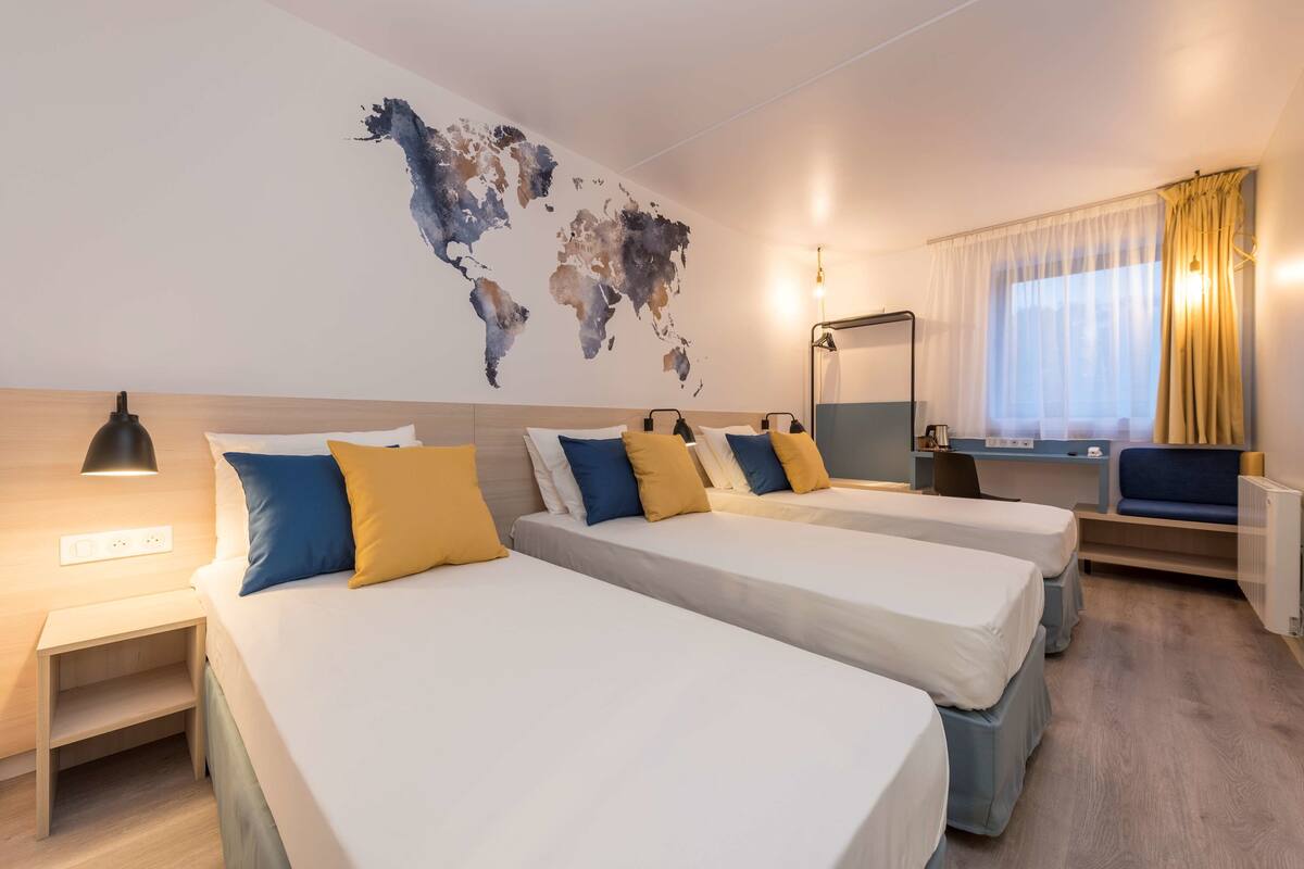 Une chambre d’hôtel avec trois lits, une carte du monde accrochée au mur, un bureau et une fenêtre avec des rideaux.