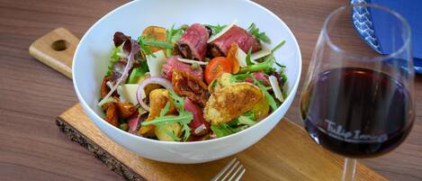 Een saladekom met diverse ingrediënten en een glas rode wijn op een houten tafel.