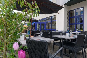 Un área de comedor al aire libre con mesas y sillas, un paraguas grande y un edificio con ventanas de marco azul al fondo.
