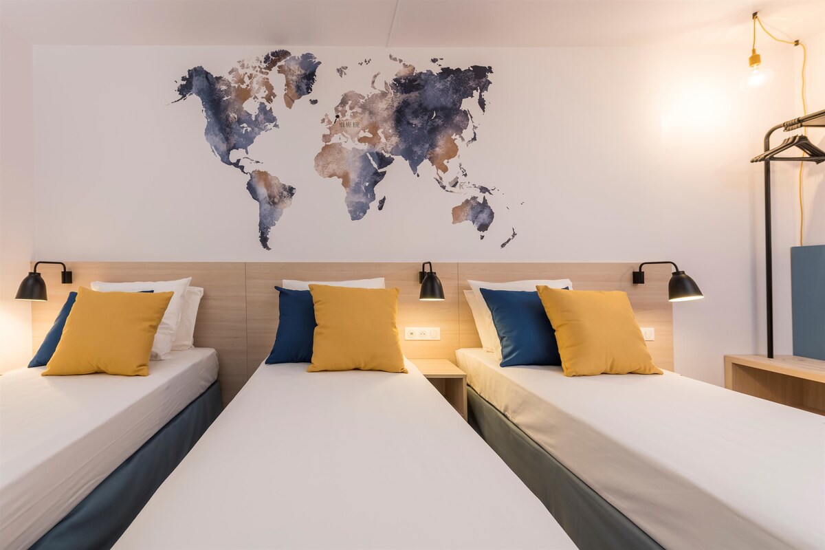 A hotel room with two beds, each with white linens and yellow and blue pillows. There is a world map on the wall and a lamp on each side of the bed.