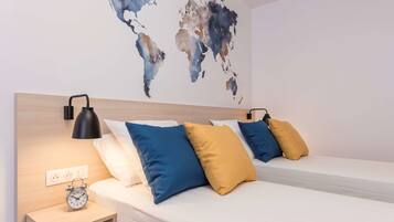 A hotel room with two beds, a world map on the wall, a bedside table with an alarm clock, and a lamp.