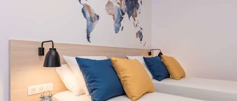 A hotel room with two beds, a world map on the wall, a bedside table with an alarm clock, and a lamp.
