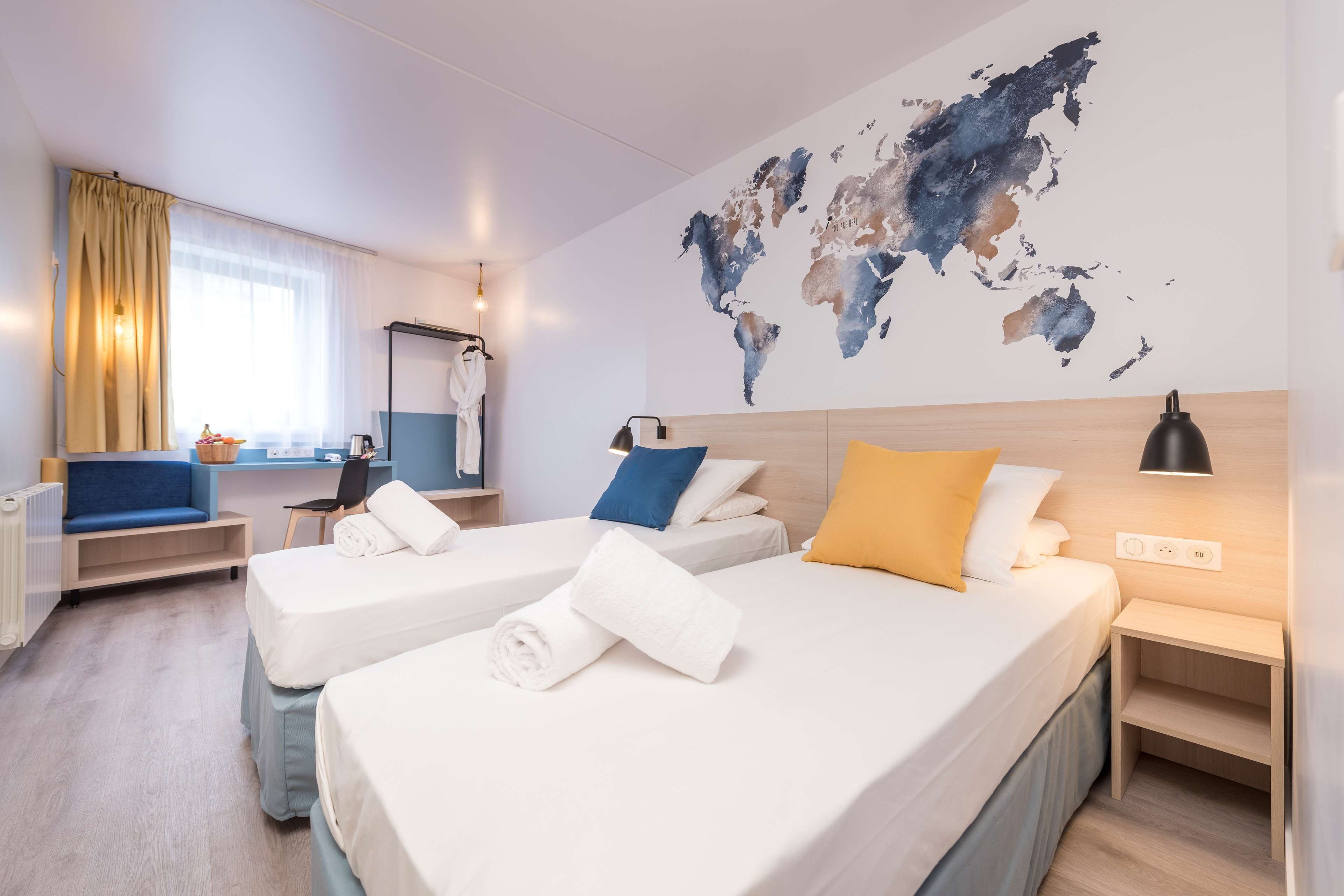 Une chambre d’hôtel avec deux lits, une carte du monde accrochée au mur, un bureau et une fenêtre avec des rideaux.