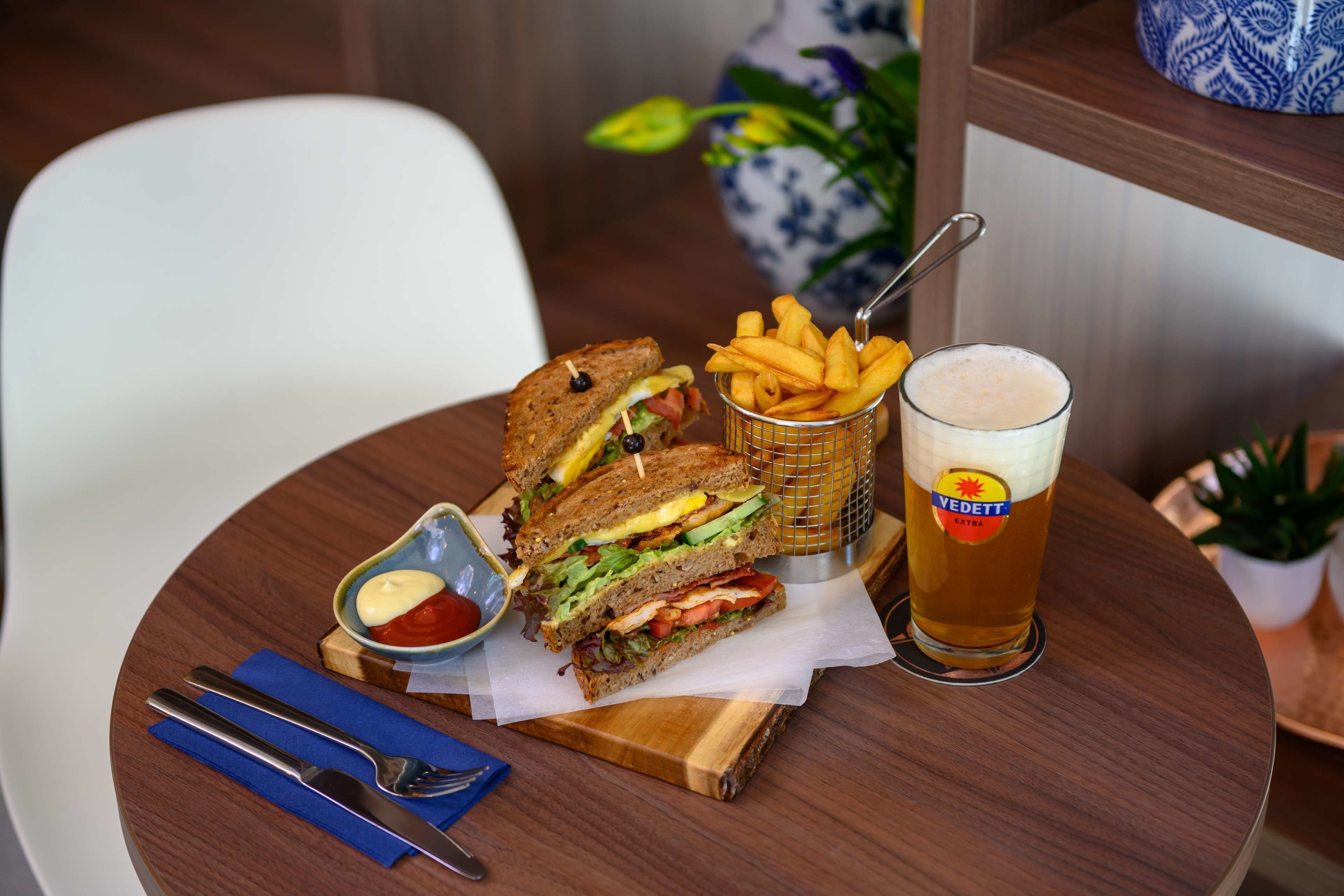 A meal consisting of a sandwich, fries, a small bowl of ketchup, and a glass of beer on a wooden table.