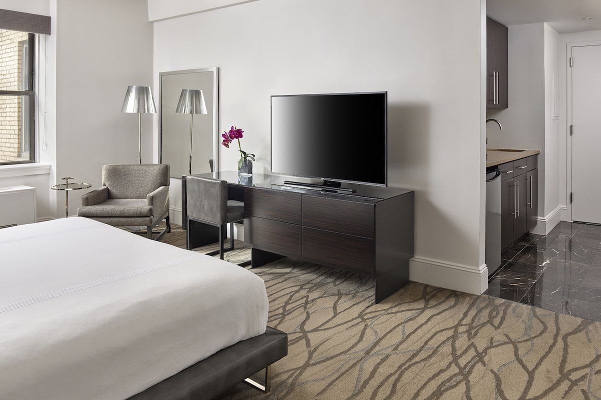 A modern hotel room with a bed, a desk with a flat-screen TV, a chair, a lamp, and a small table.