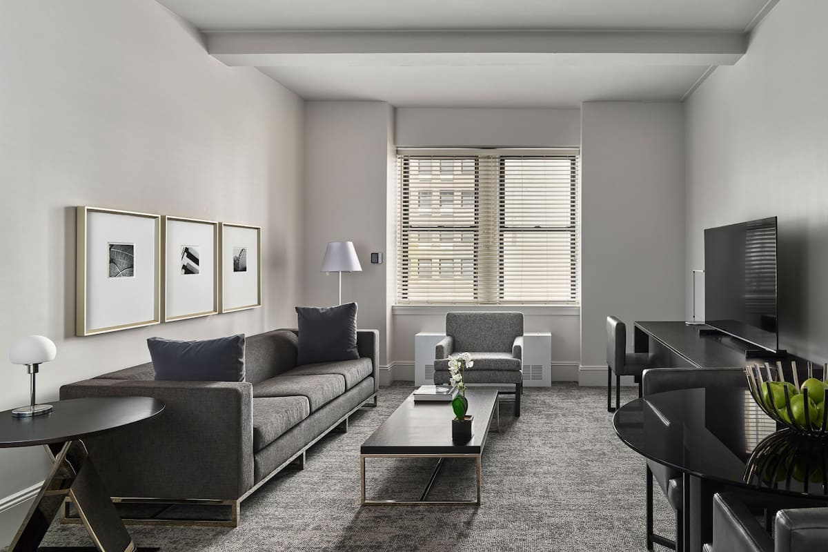 A modern living room with a sofa, coffee table, and TV. There are framed pictures on the wall and a window with blinds.