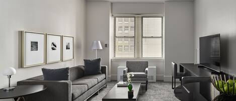 A modern living room with a sofa, coffee table, and TV. There are framed pictures on the wall and a window with blinds.