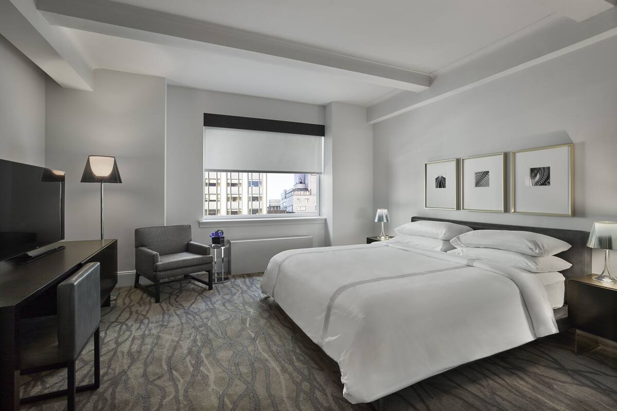 A modern hotel room with a large bed, a desk, a chair, a TV, and framed artwork on the wall.