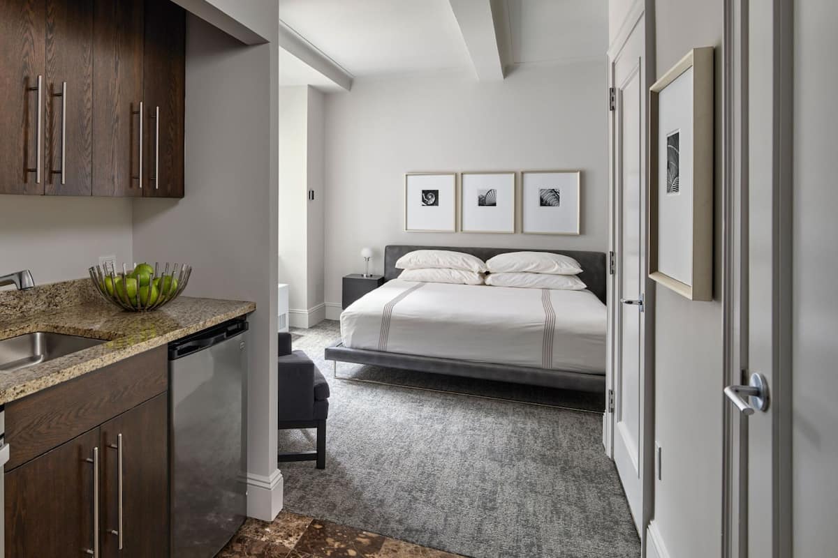 A modern bedroom with a bed, bedside table, and a nightstand. There is a kitchen area with a sink, dishwasher, and a fruit bowl. The room has a neutral color palette and a carpeted floor.