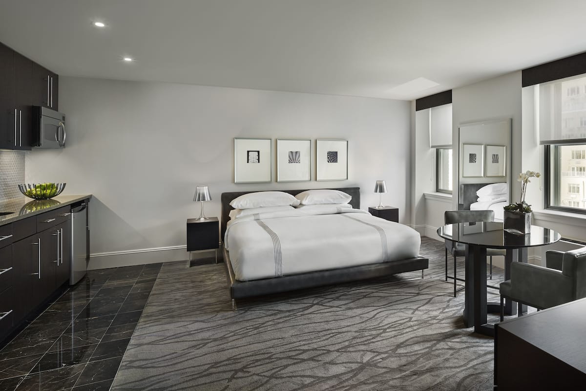 A modern hotel room with a large bed, a dining table, a small kitchen area, and artwork on the walls.