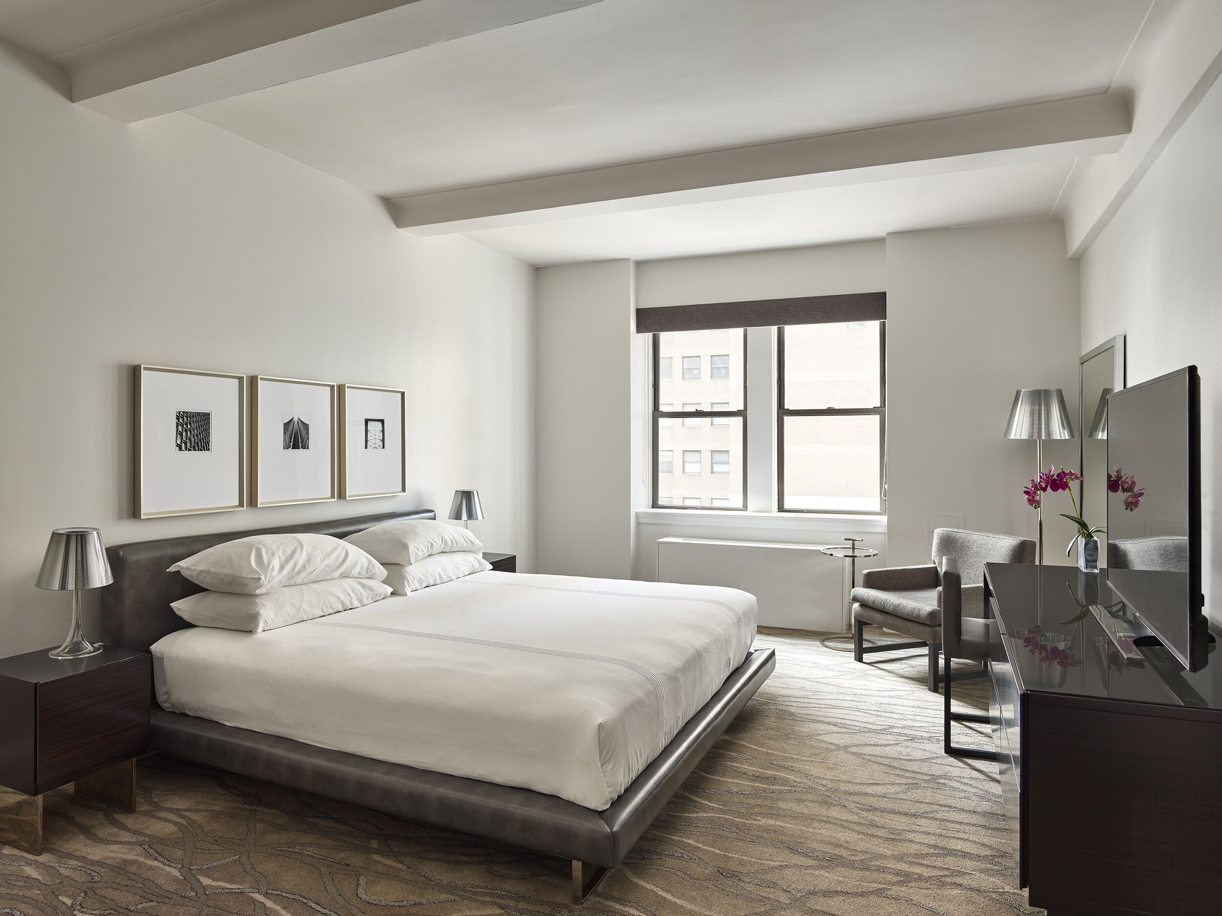 A modern hotel room with a large bed, a desk, a chair, a window with a view of buildings, and framed pictures on the wall.