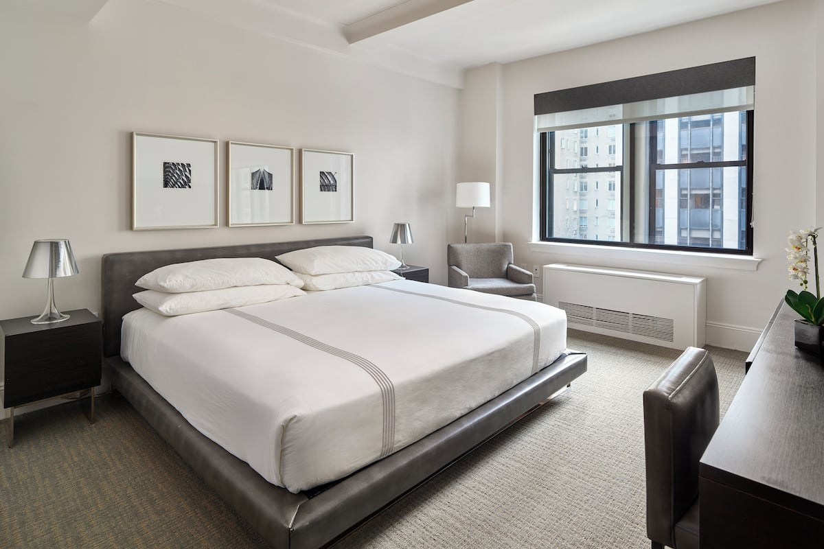 A modern bedroom with a large bed, a desk, a chair, a radiator, and framed artwork on the wall.