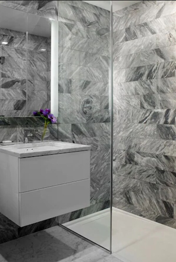 Une salle de bain moderne avec un pare-feu en marbre, une douche en verre et un meuble-lavabo blanc agrémenté d’un vase à fleurs violet.