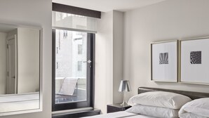 A hotel room with a large bed, a nightstand with a lamp, a window with a view of buildings, and two framed pictures on the wall. - AKA Central Park (New York)