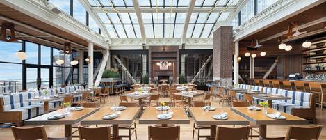 En rummelig restaurant med højt til loftet, store vinduer og en central pejs. Bordene er pænt anrettet med stole, og spiseområdet er godt oplyst.