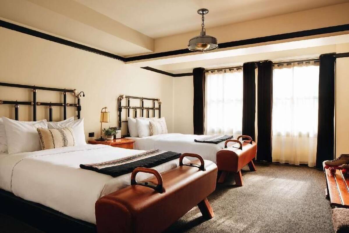 A hotel room with two beds, a bench, a piano, and a large window with curtains.