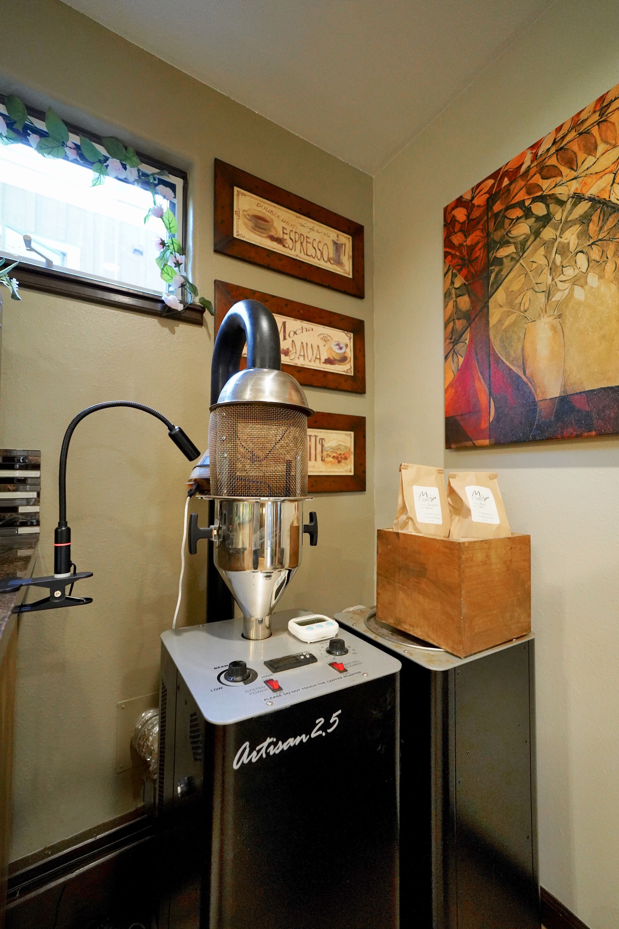 A coffee machine with a metal body and a control panel, a window with a decorative vine, and framed certificates on the wall.