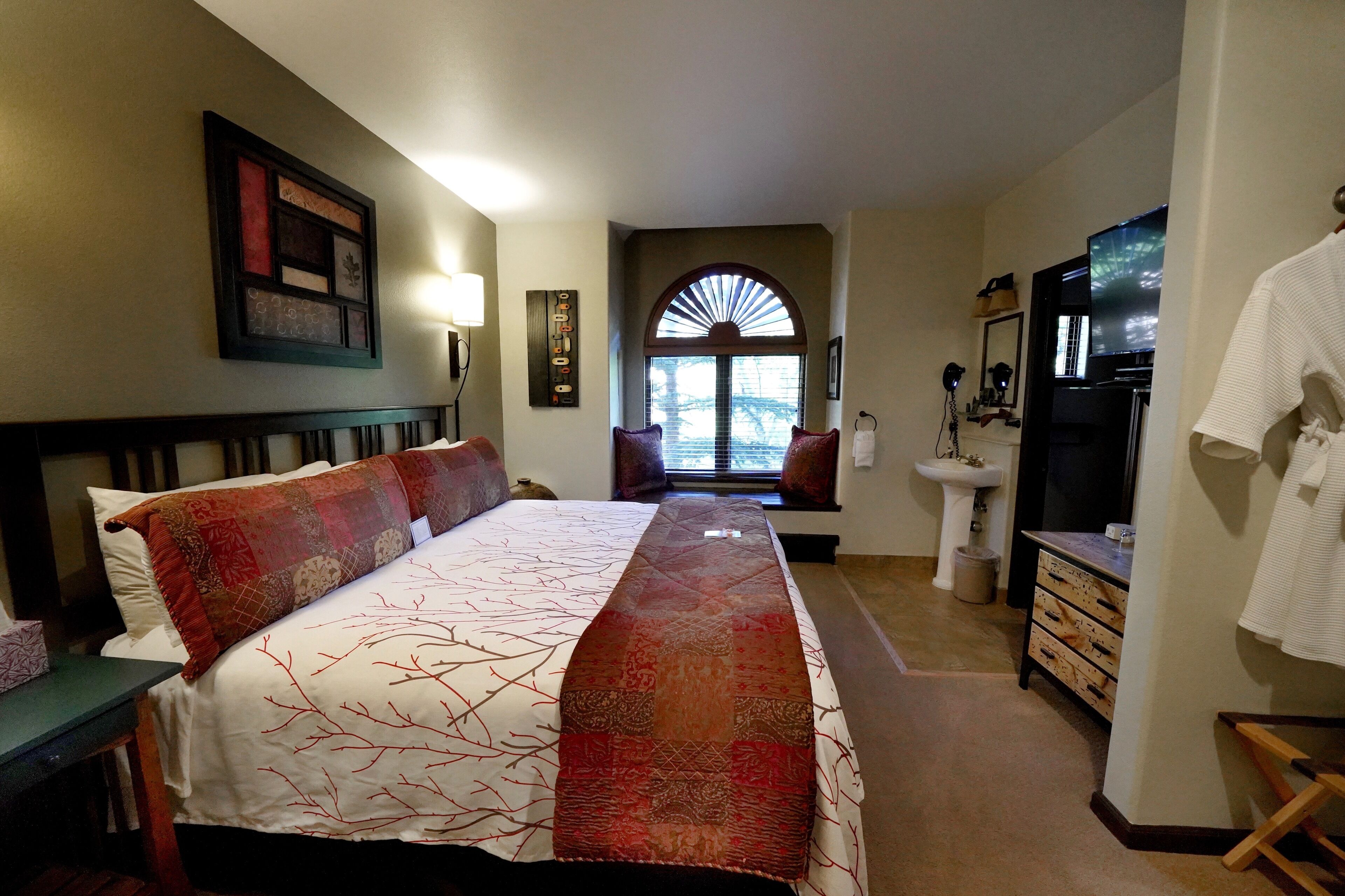 A hotel room with a large bed, a vanity with a mirror, a sink, and a window with a view of trees.