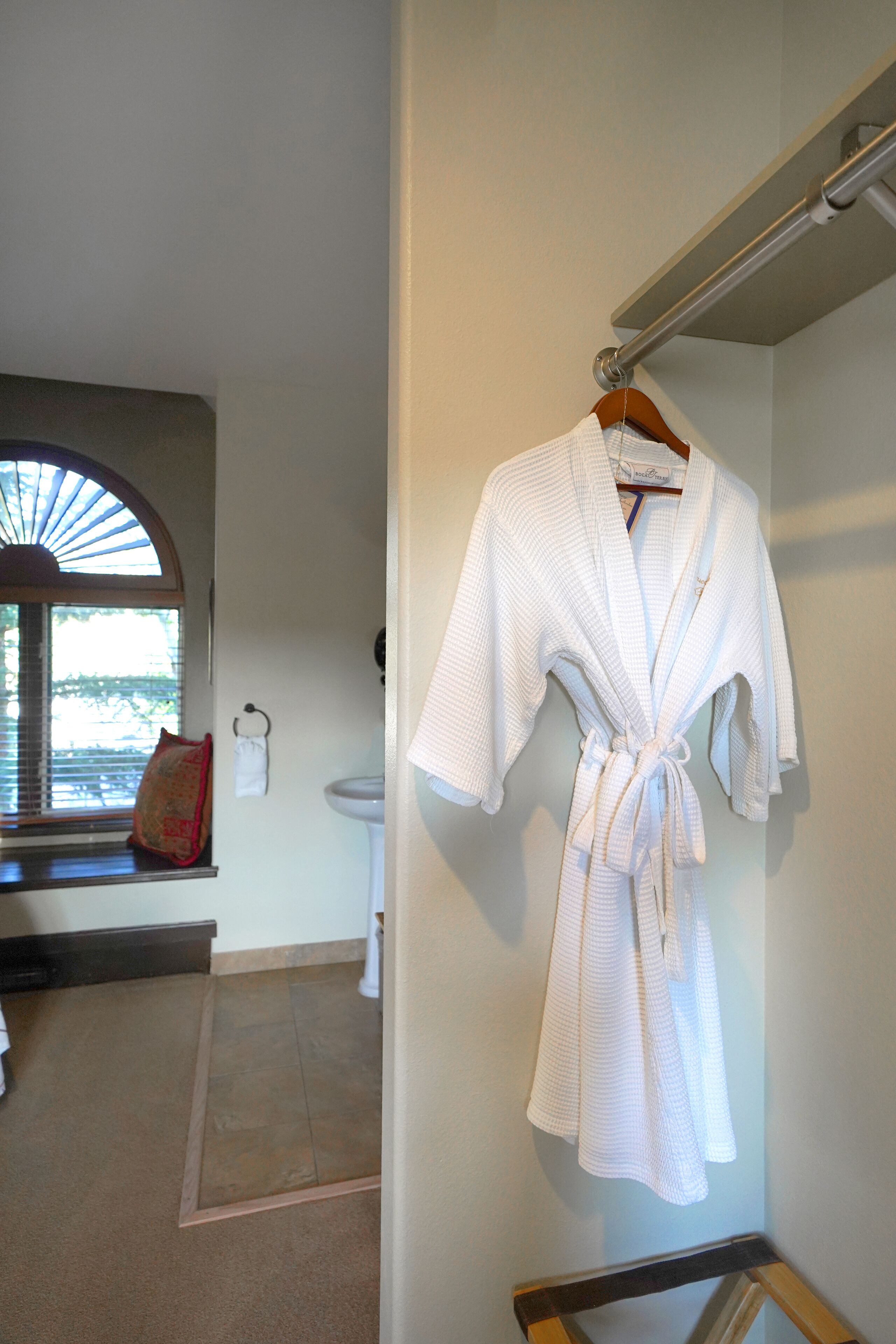 A bathroom with a white robe hanging on a rack, a window with blinds, a towel rack, and a sink.
