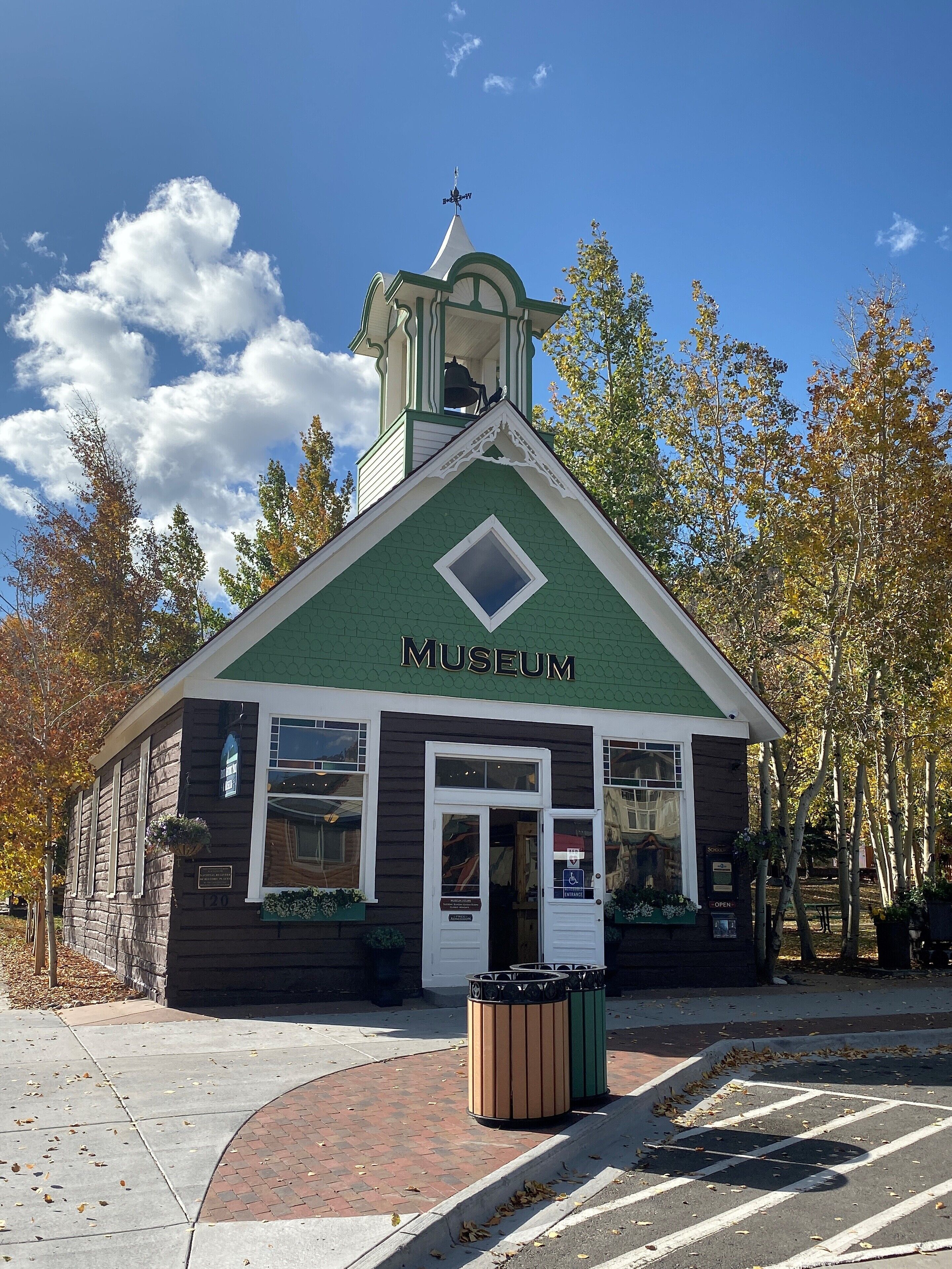 A small museum building with a green roof and a bell tower, surrounded by trees with autumn leaves.