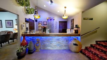 Zona de recepción de hotel con mostrador de piedra, dos lámparas colgantes y un cartel que indica un spa y una sala de juegos.