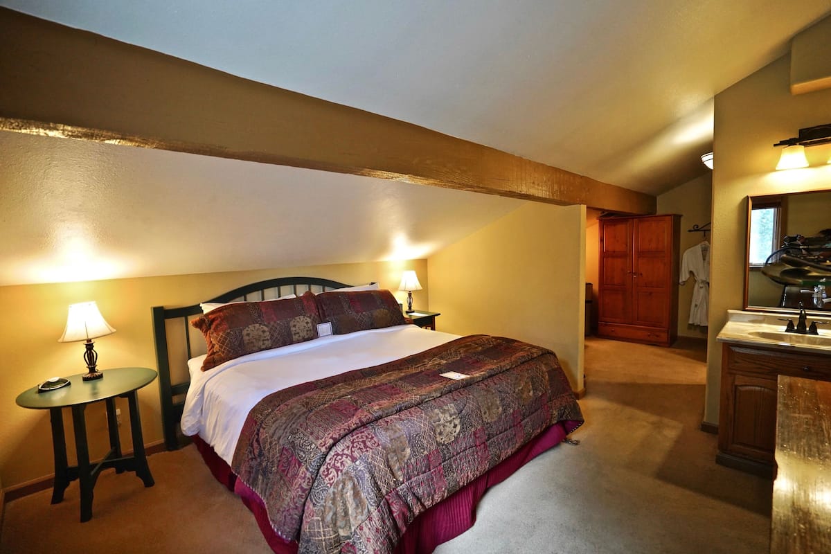 A bedroom with a bed, a nightstand with a lamp, a wooden wardrobe, and a bathroom with a mirror and sink.