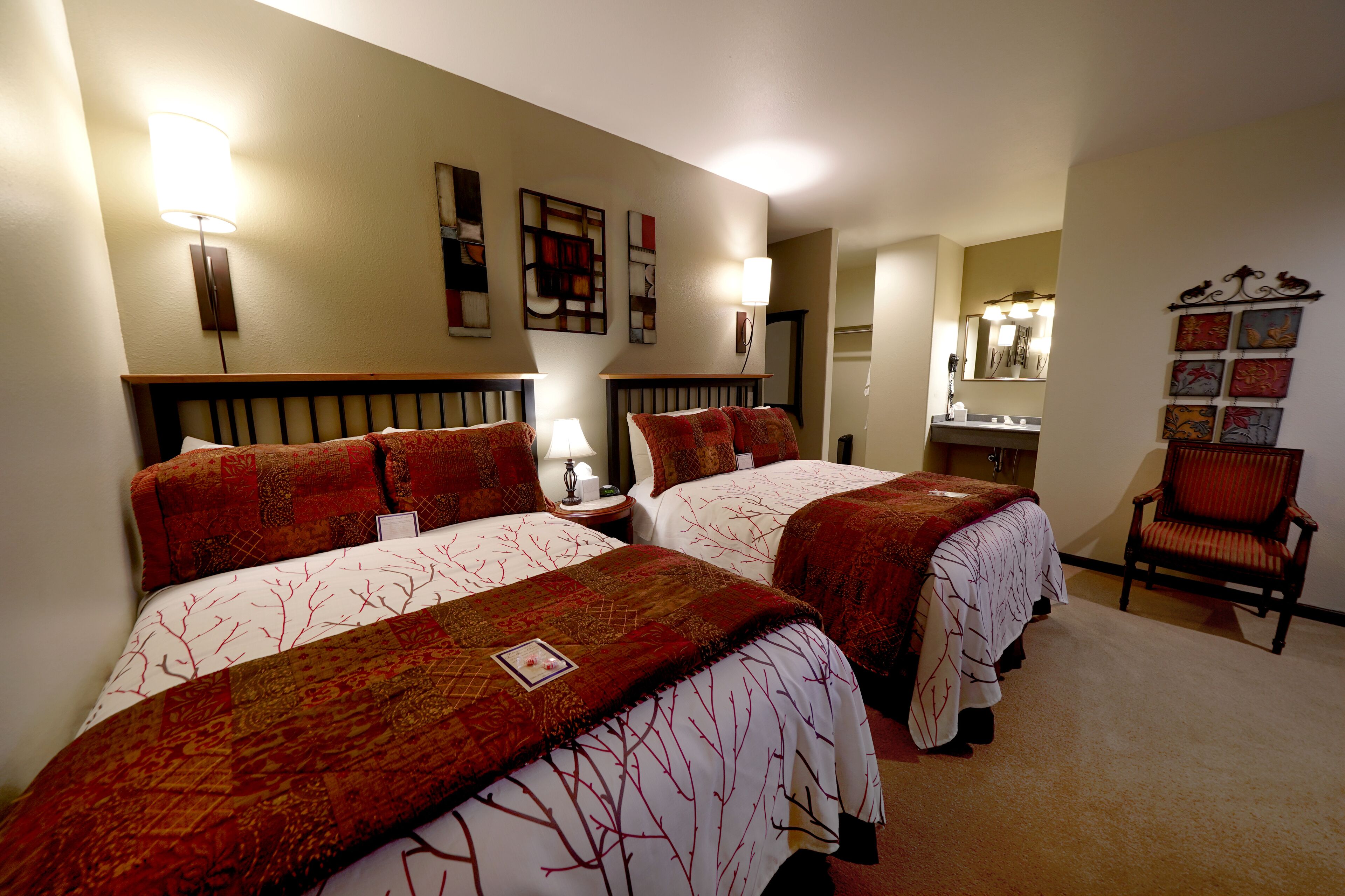 A hotel room with two beds, a sitting area with a chair, a vanity with a mirror, and wall art.