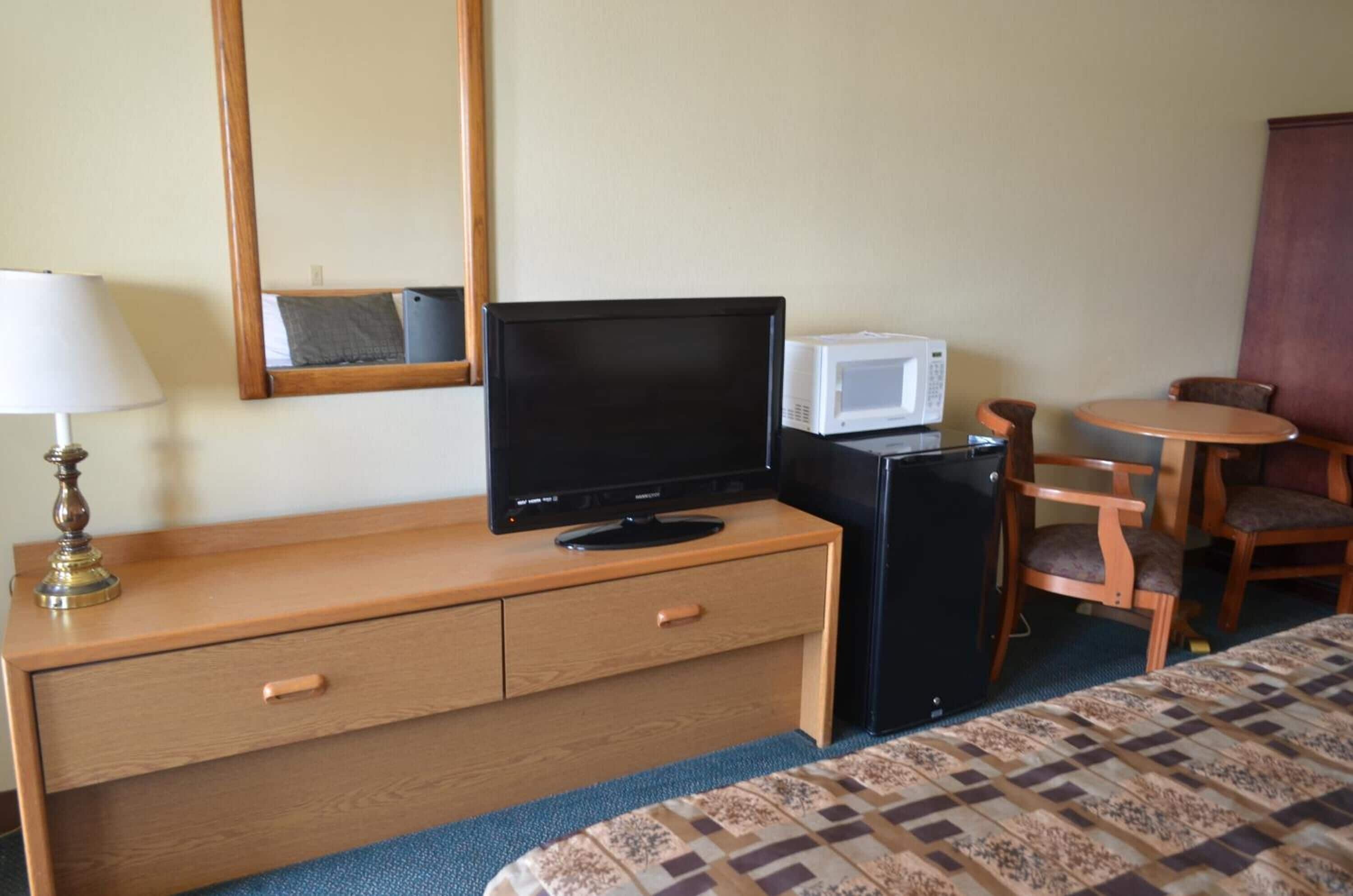 Une chambre d’hôtel comprenant un lit, une commode en bois avec des tiroirs, une télévision, un four à micro-ondes, un petit réfrigérateur, une lampe et une table ronde avec deux chaises.