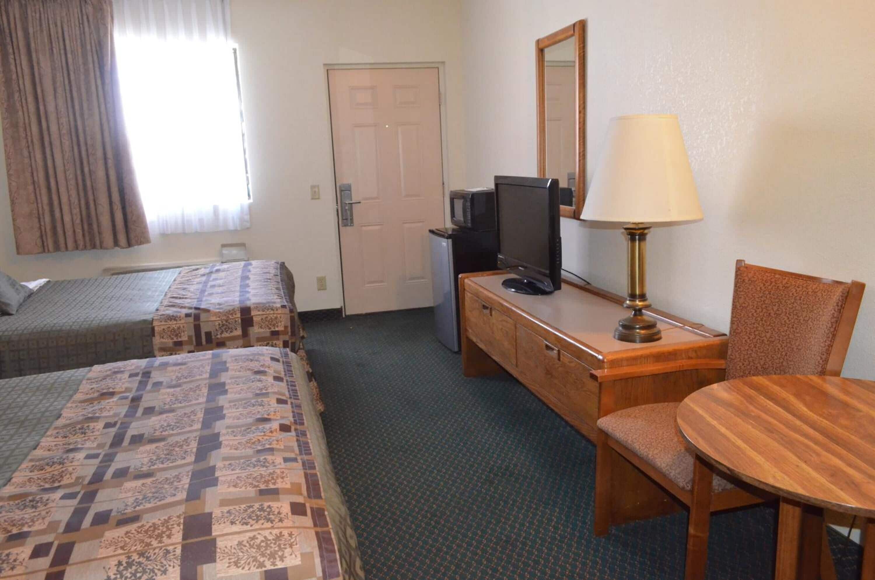Quarto de hotel com duas camas de solteiro, uma mesa com computador e luminária, uma cadeira e uma pequena mesa.