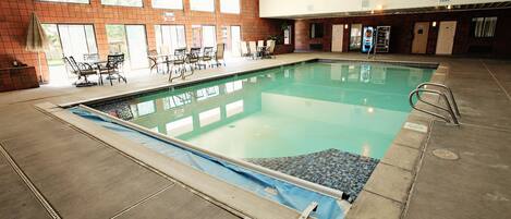 An indoor swimming pool with tiled walls, a blue pool deck, and a seating area with tables and chairs.