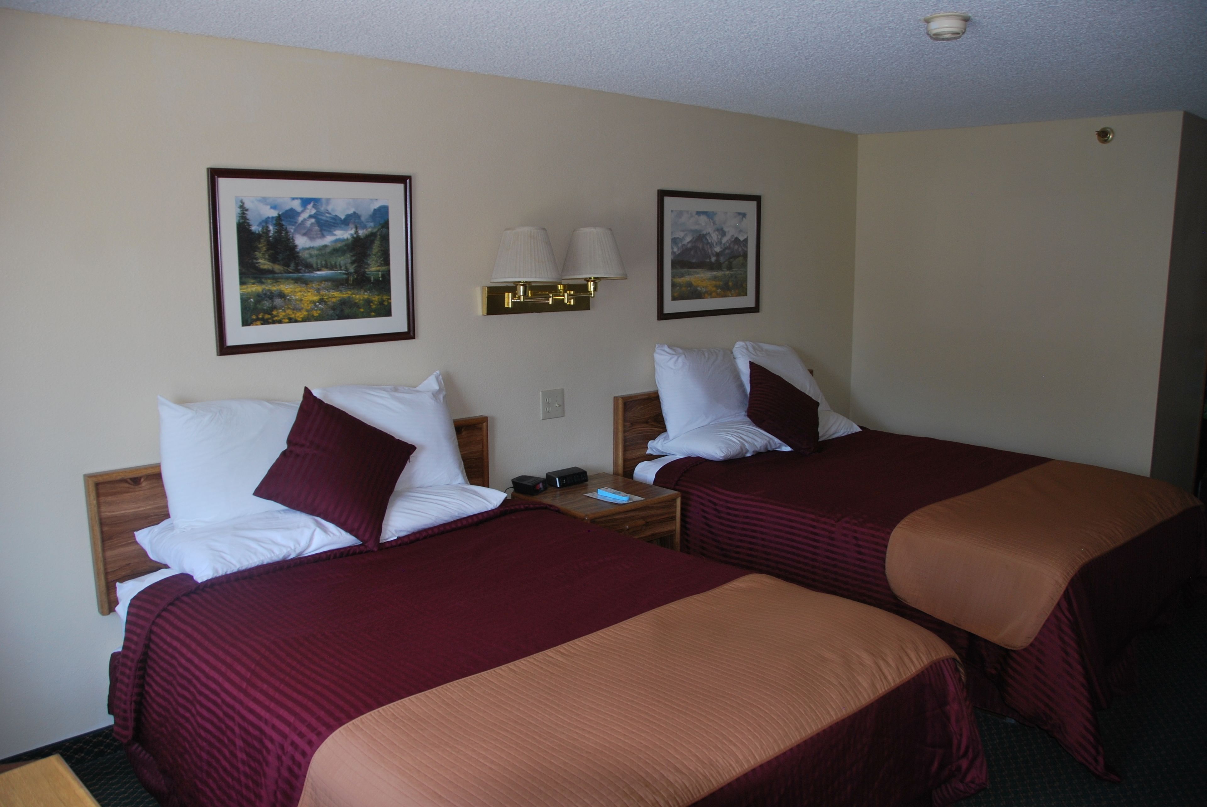 Quarto de hotel com duas camas, cada uma com colcha vermelha e branca, mesa de cabeceira com abajur e duas fotos emolduradas na parede.