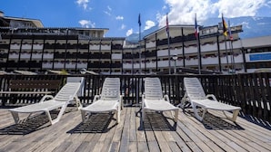 A wooden deck with white lounge chairs, a building with balconies, and flags in the background.