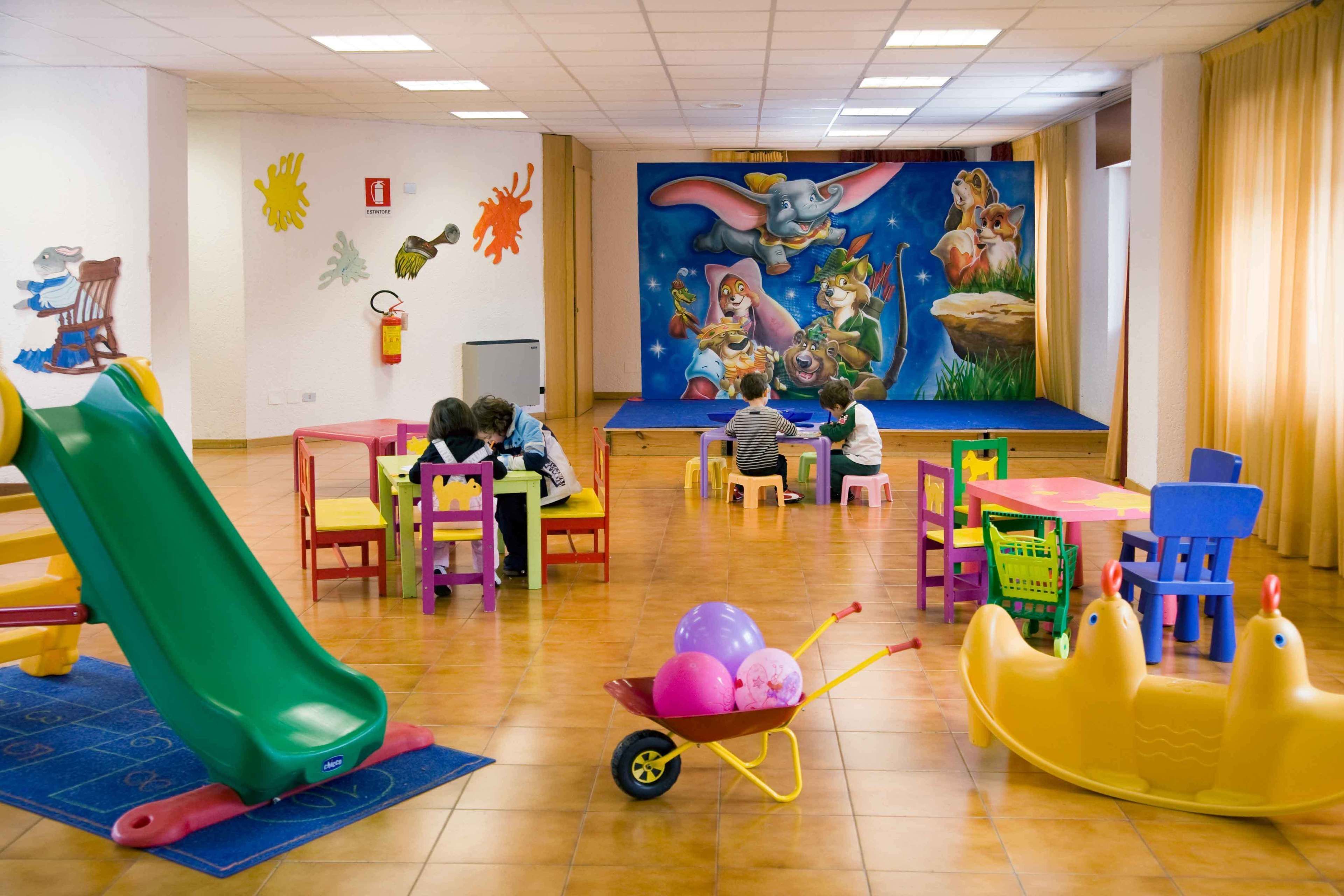 A children's playroom with a slide, colorful chairs, tables, and a mural on the wall.