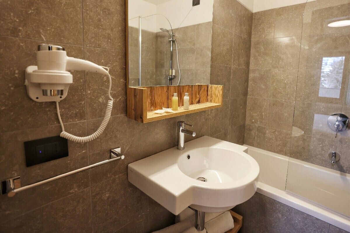Et moderne badeværelse med en hvid vask, en træhylde med toiletartikler, en hårtørrer og en brusekabine med glasvægge.