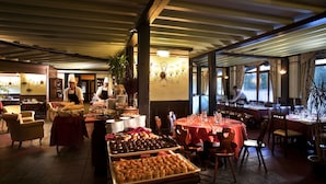 A restaurant with multiple tables set for dining, a buffet with various pastries, and a kitchen area with chefs preparing food.