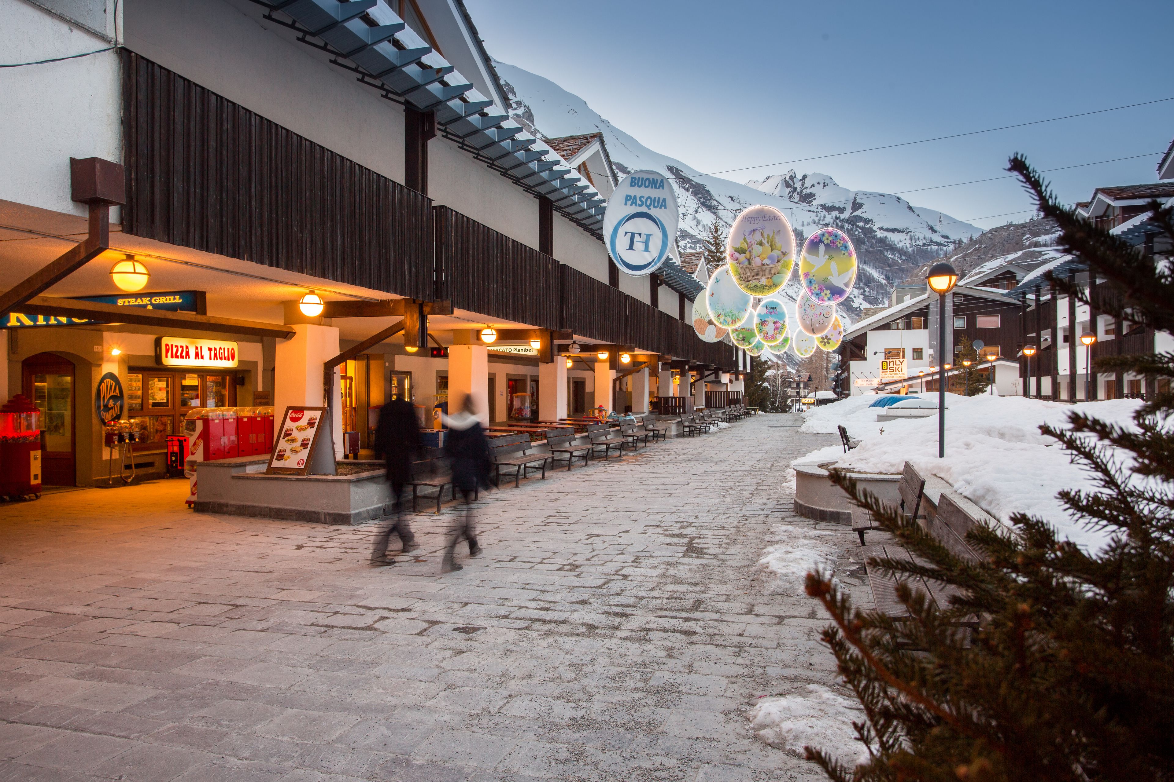 A snow-covered street with a restaurant named "PIZZA AL TAGIO" and a sign for "BUONA PASQUA".