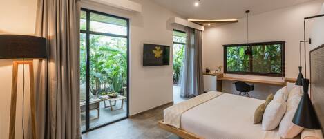 A modern bedroom with a large bed, a desk, and a TV. There is a sliding glass door leading to a balcony with plants.