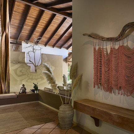 A room with a wooden ceiling, a bench, a wall-mounted macrame piece, and a vase with pampas grass.