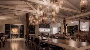 A spacious dining area with a long wooden table and chairs, hanging wicker chandeliers, and a wine rack in the background.