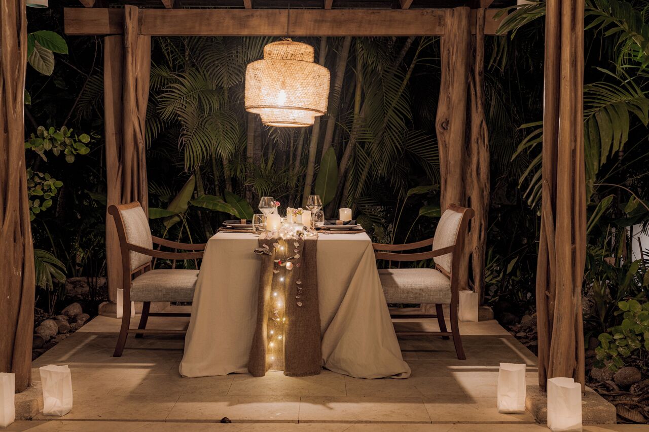 An outdoor dining area with a table set for two, surrounded by wooden chairs, under a pergola with a woven light fixture.