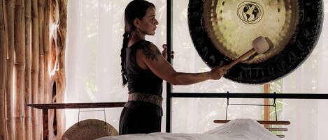 A person performing a sound therapy session with a large gong in a room with bamboo walls and a bed.