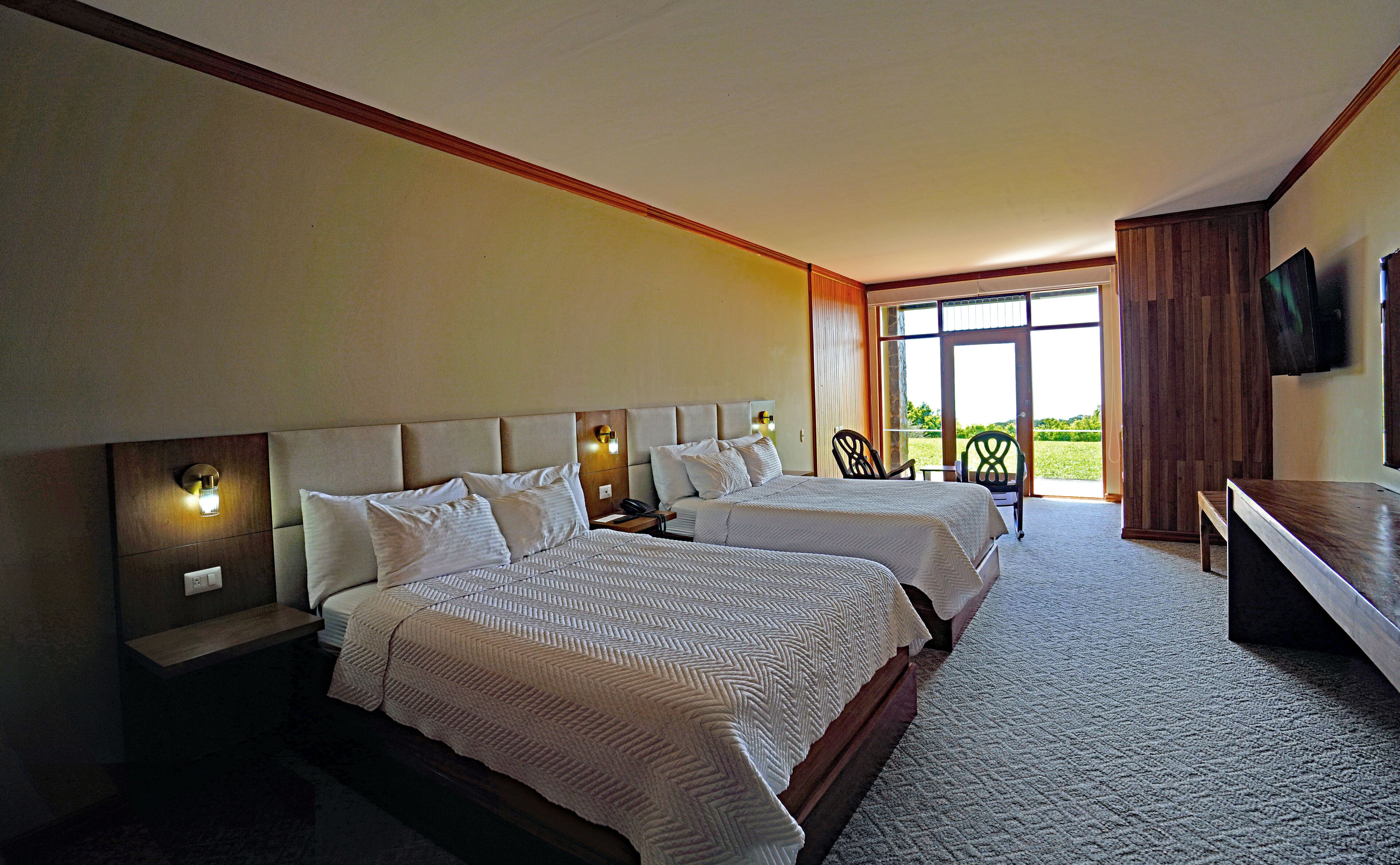 A hotel room with two beds, a desk, a chair, and a large window offering a view of the outdoors.
