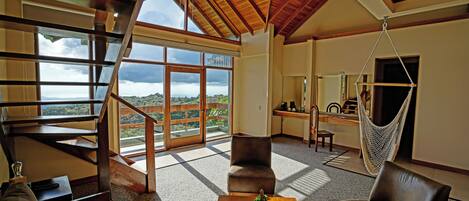 A spacious living area with a high ceiling, a wooden staircase, a hammock, and a view of the outdoors.