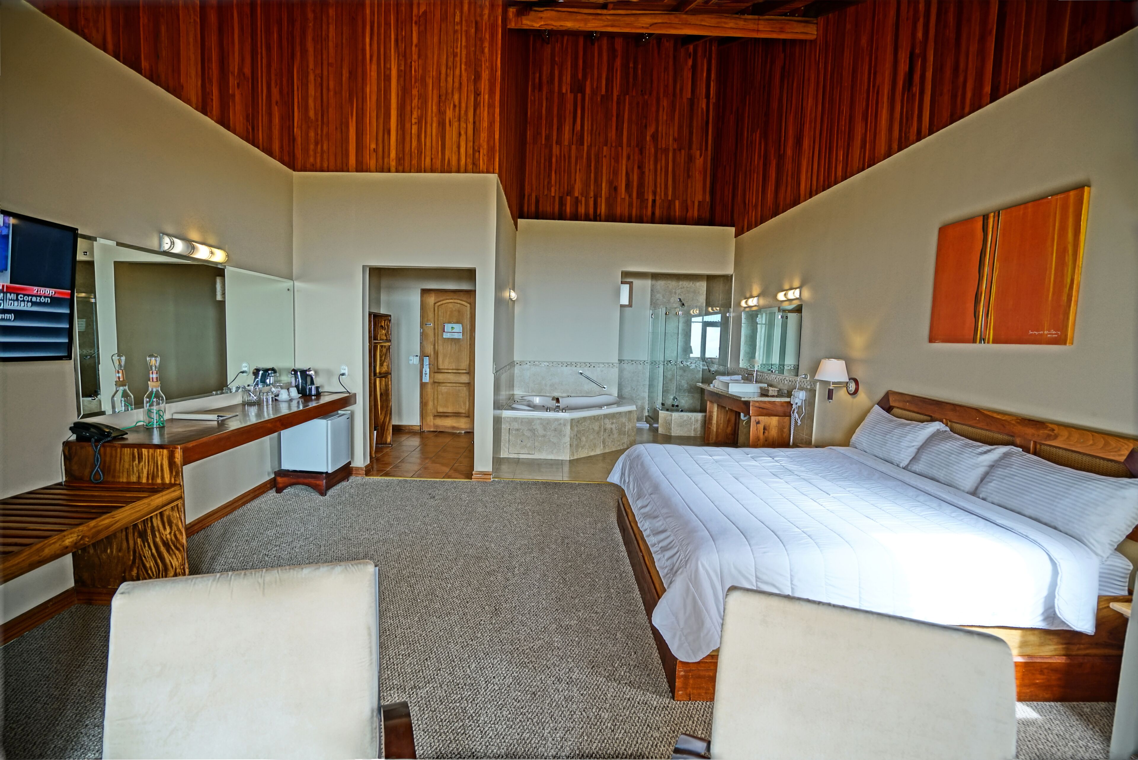 Une chambre d’hôtel avec un plafond en bois, un grand lit, un bureau avec un miroir, une salle de bain avec une baignoire et un petit coin salon.
