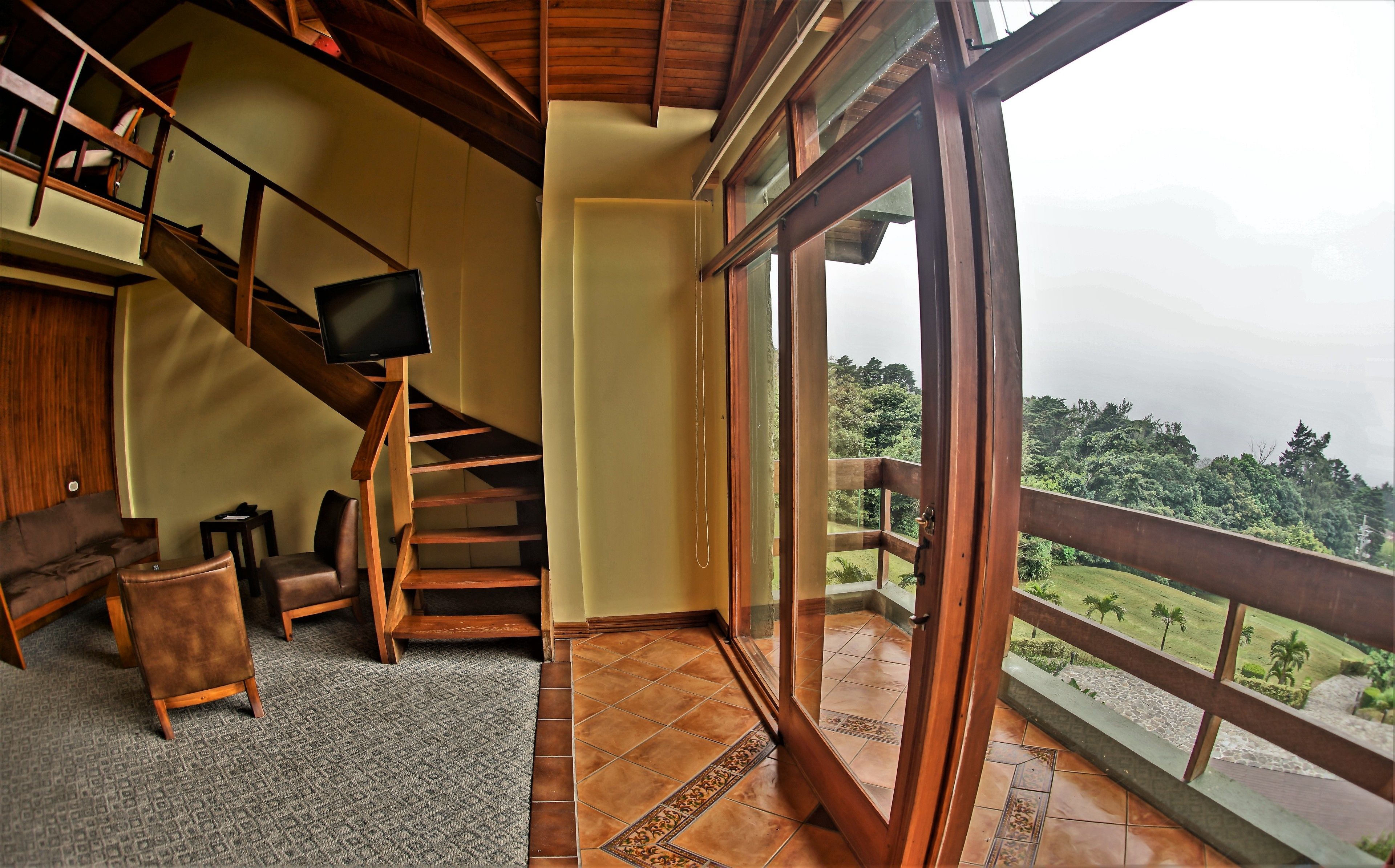 A spacious room with a wooden staircase, a large window with a view of a lush landscape, and a cozy seating area with a sofa and armchairs.