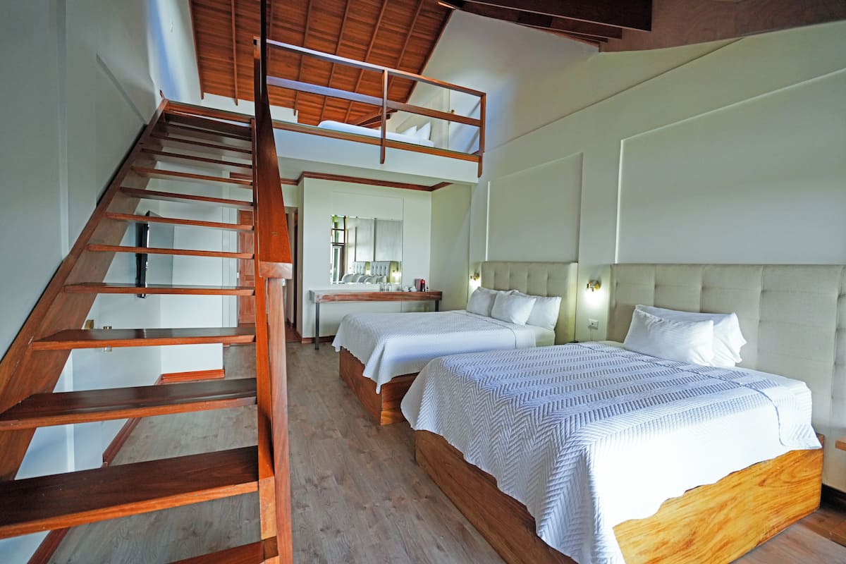 Une chambre d’hôtel avec un escalier en bois, deux lits avec du linge de lit blanc, un coin cuisine à l’arrière-plan et une grande fenêtre.