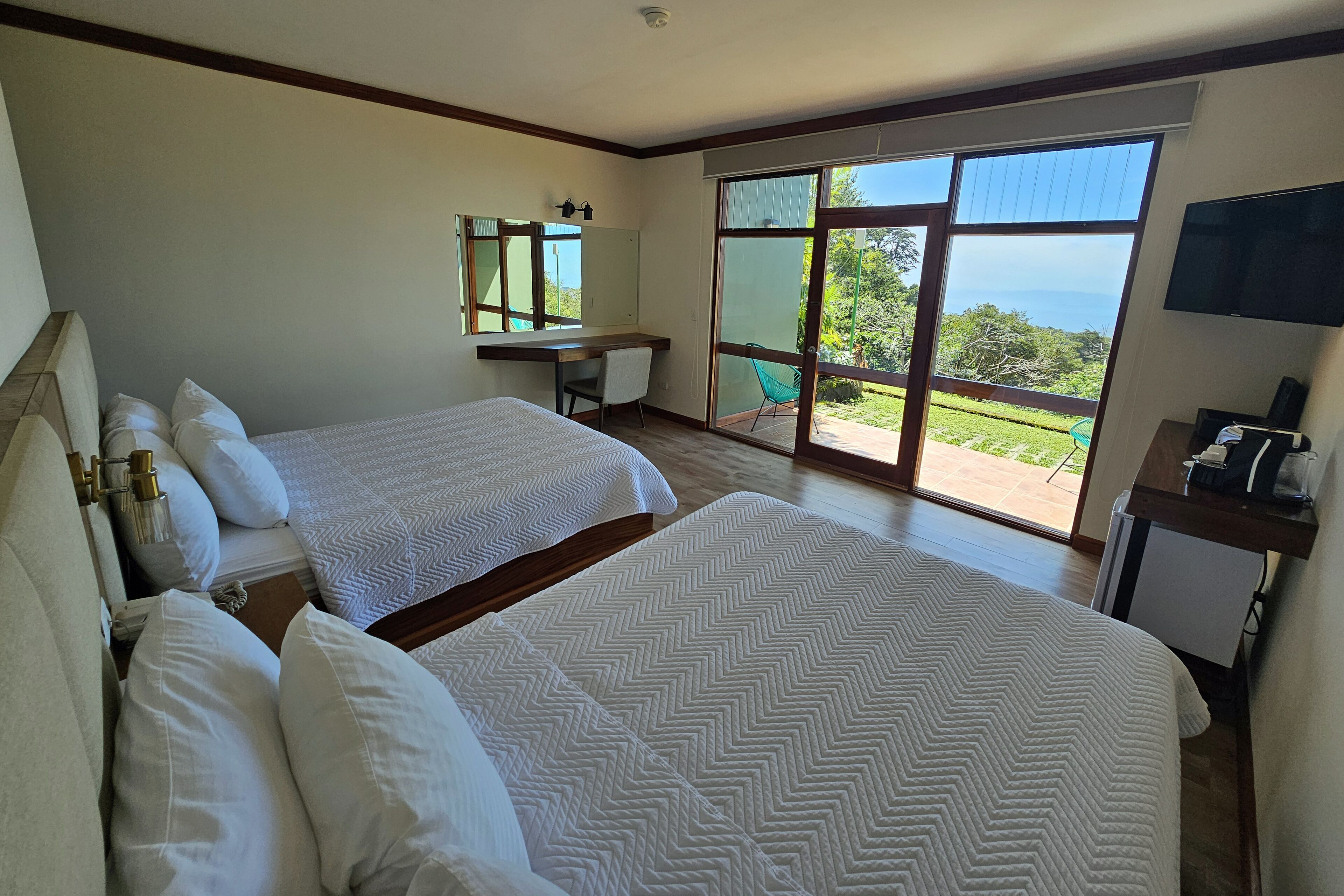 A bedroom with two beds, a desk, a chair, a TV, and a balcony with a view.